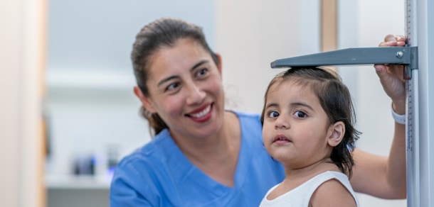 A nurse in a medical office is measuring the height of a young child. The nurse is wearing blue hospital scrubs; the child wears a white dress. The nurse is looking at the measuring device; the child looks at the camera. Both are smiling.
