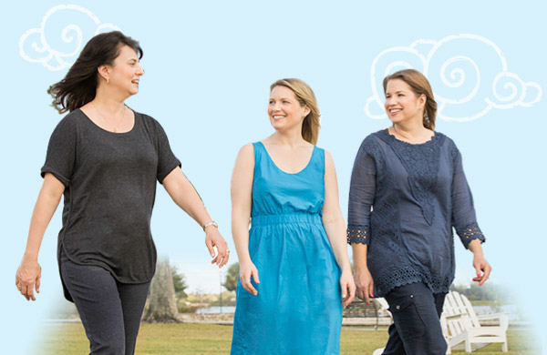 three women outdoors walking toward the camera, looking at each other and smiling with trees in the background.