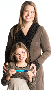 three-quarters view of Shauna standing behind her daughter Aria, a Norditropin patient, with her hands on Aria&rsquo;s shoulders while Aria holds a Flexpro pen with both hands.