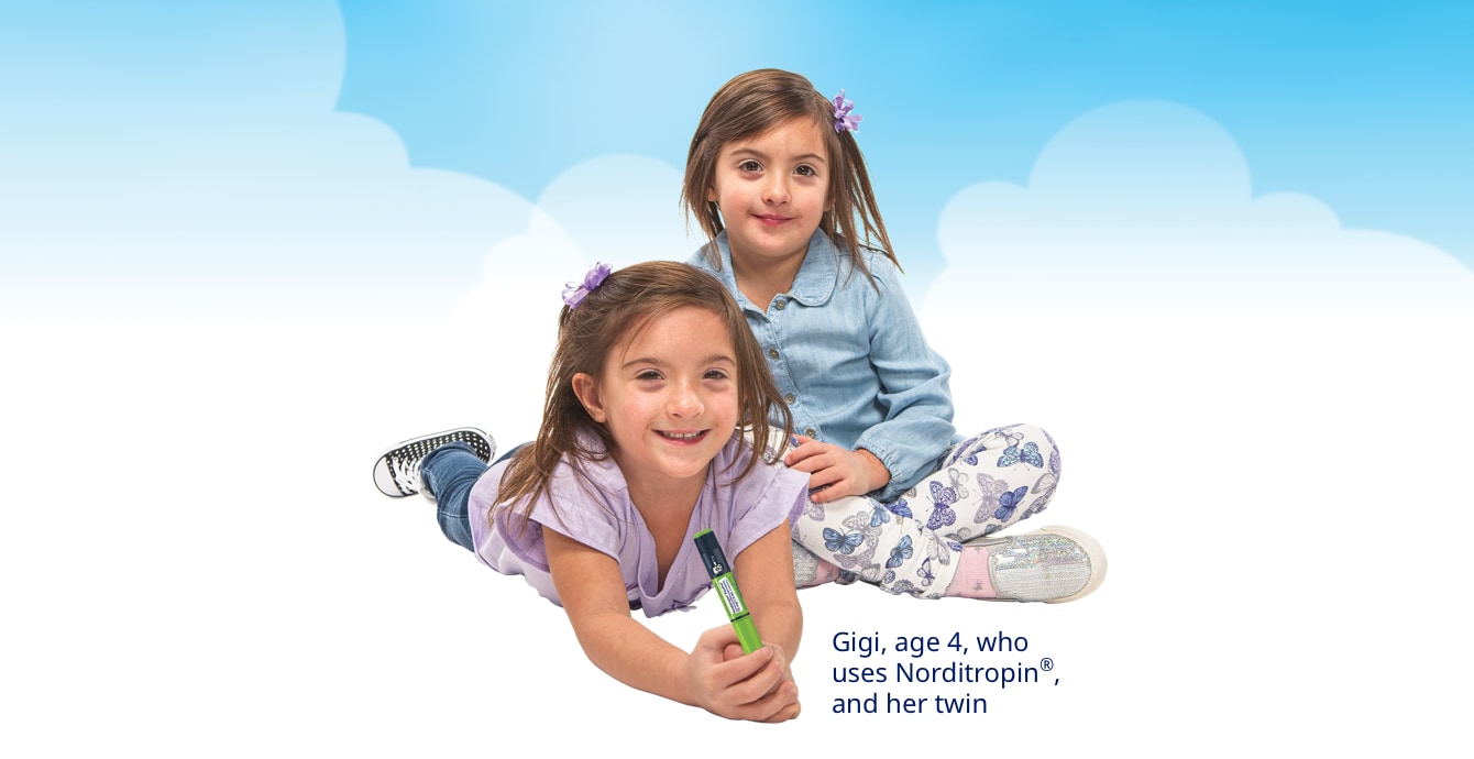 Two young girls who appear to be twins are sitting close to each other and smiling. They have long brown hair and are wearing play clothes in various shades of blue. Both are wearing sneakers. One is holding a FlexPro® pen used for injections.