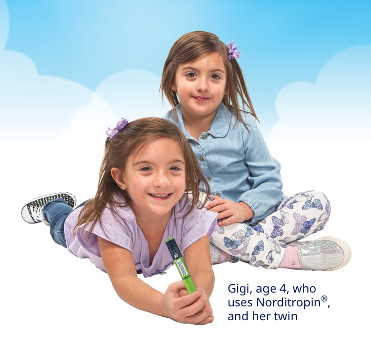 Two young girls who appear to be twins are sitting close to each other and smiling. They have long brown hair and are wearing play clothes in various shades of blue. Both are wearing sneakers. One is holding a FlexPro® pen used for injections.