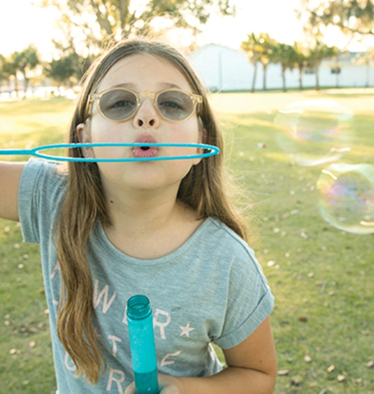 Aria, an 8-year-old girl, is pictured standing outside. She is wearing a gray T-shirt and glasses and holding a blue bubble wand. She is blowing bubbles that float off to the right. Behind her is a green field, many trees, an indistinct white building, and sunlight.