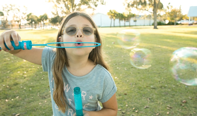 Aria, an 8-year-old girl, is pictured standing outside. She is wearing a gray T-shirt and glasses and holding a blue bubble wand. She is blowing bubbles that float off to the right. Behind her is a green field, many trees, an indistinct white building, and sunlight.