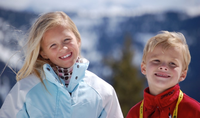 A 10-year-old girl, Logan, stands outside next to her brother. Both are wearing cold-weather coats. Logan has blonde hair and is tilting her head to the side; she is also wearing a scarf. Her brother also has blonde hair; he has a yellow strap visible over his shoulders. They are smiling. Behind them is a blurry mountain scene.