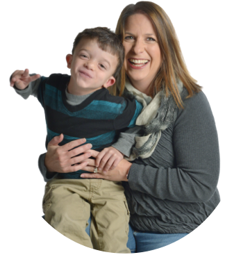 A mother, Amanda, holds her son, Colton, age 5, on her lap. Colton is flashing a ‘peace’ sign and has tilted his head; he is wearing a striped sweater and casual tan pants. Amanda is wearing a gray sweater and a scarf. Both of them are smiling.